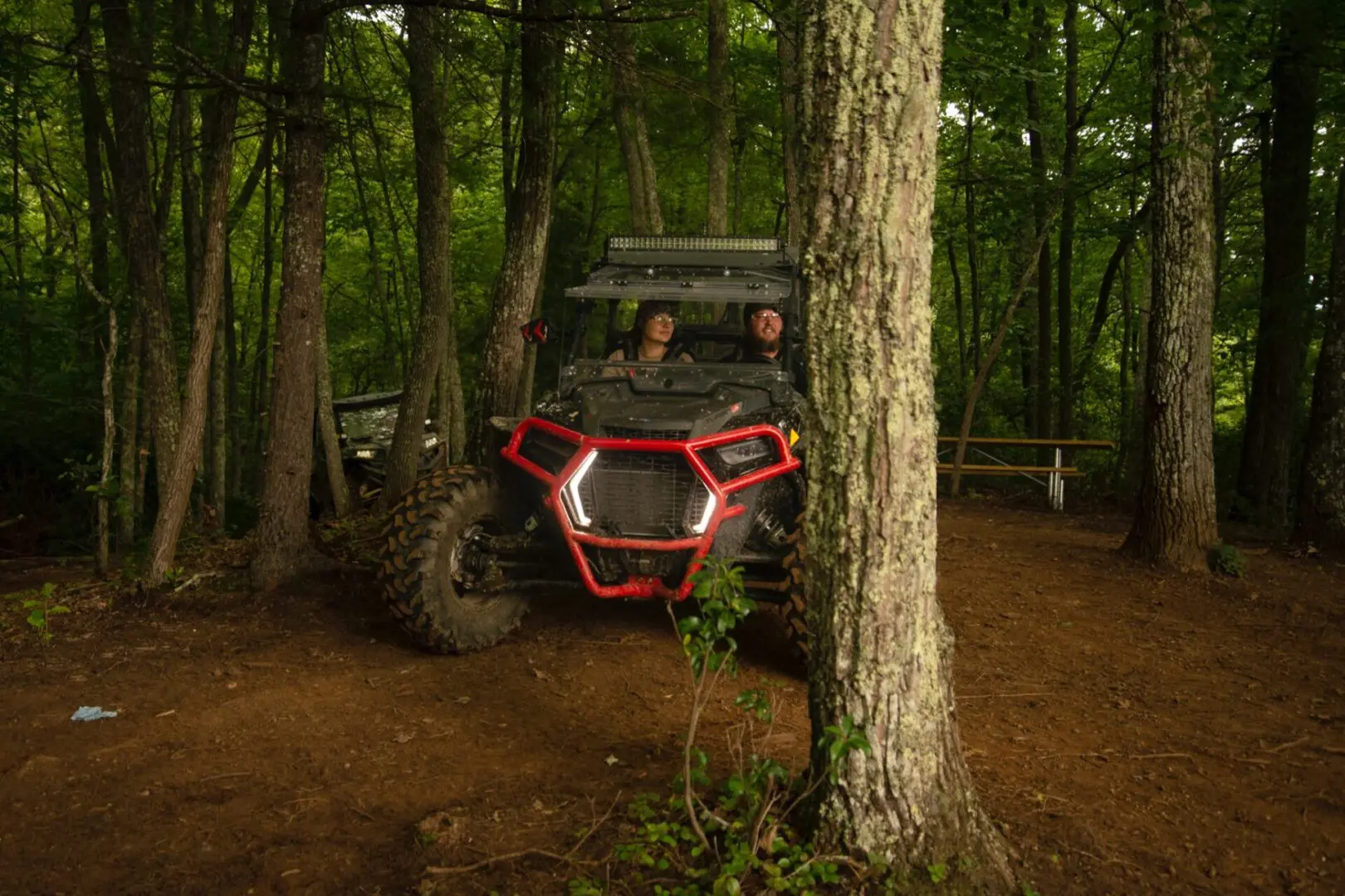Two people in a red and black vehicle parked on the side of a forest.